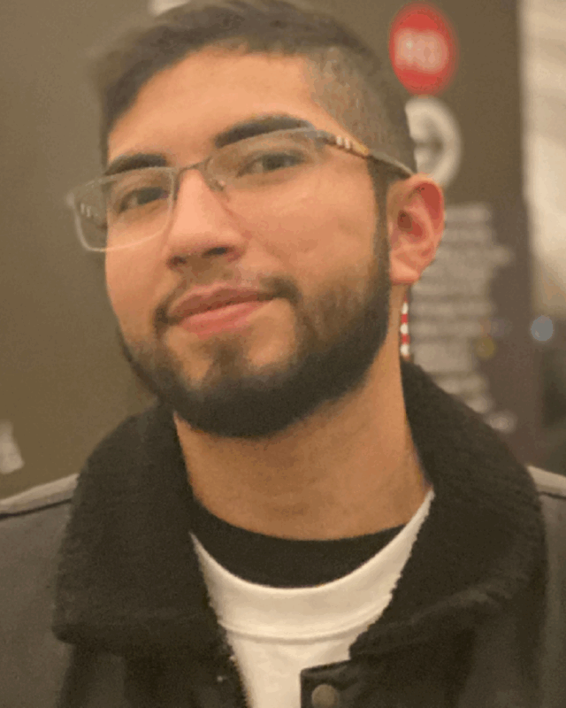 A man with a beard and glasses stands beside a train, smiling slightly as he gazes off to the side.