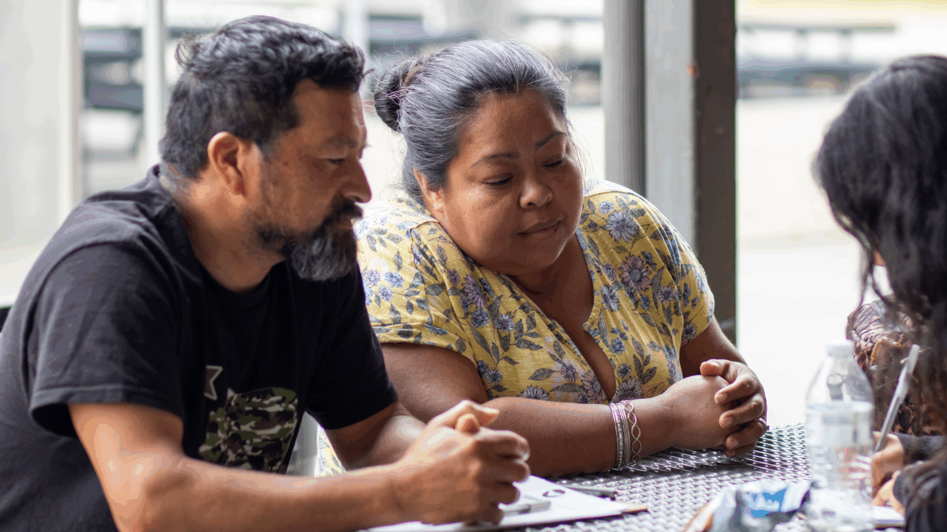Three people are seated around a table.  All three appear to be engaged in a conversation or activity.
