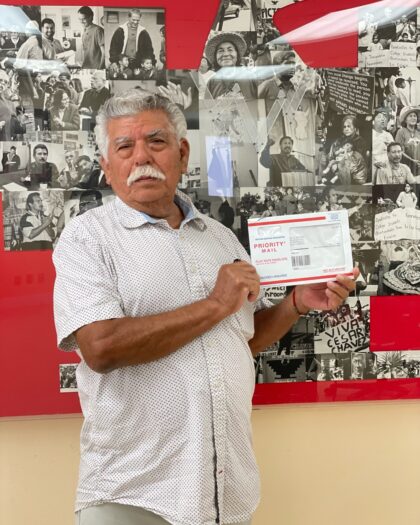 An older man with gray hair and a mustache, wearing a white short-sleeved, button-up shirt with a subtle dotted pattern.