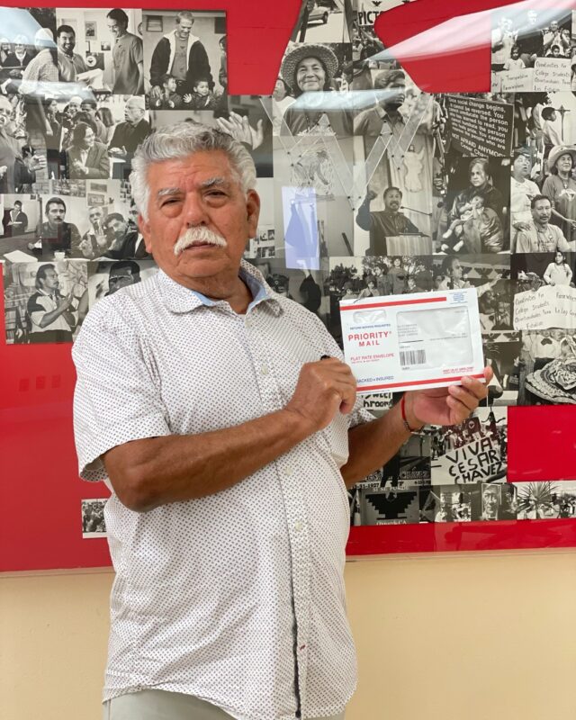 An older man with gray hair and a mustache, wearing a white short-sleeved, button-up shirt with a subtle dotted pattern.