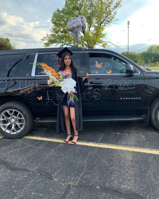 A woman wearing a graduation cap and gown is standing next to a black SUV.