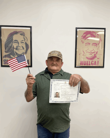 An older man holding his citizenship papers standing indoors against a plain wall with two framed posters behind him.