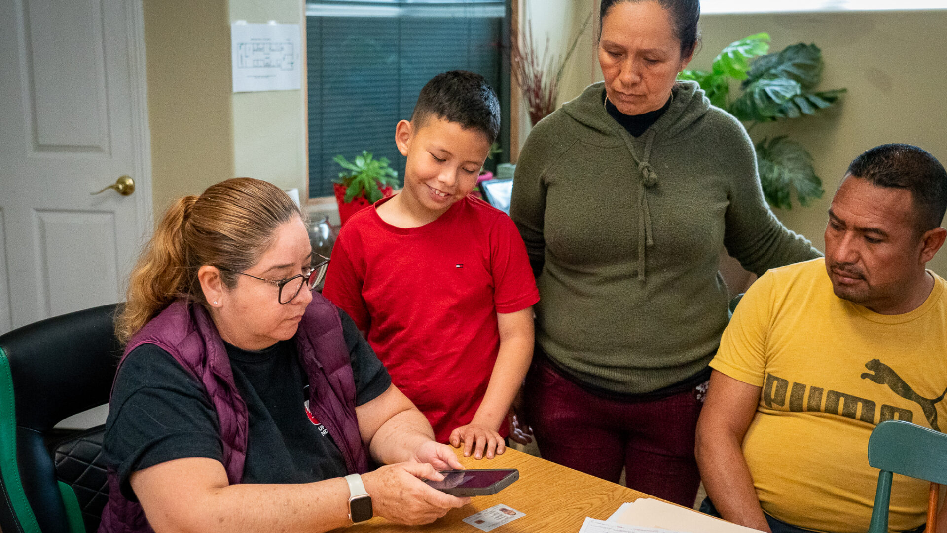 A family, consisting of two adults and a preteen child, is gathered around a light-colored wooden table. The woman, seated at the table and wearing glasses, appears to be reviewing documents