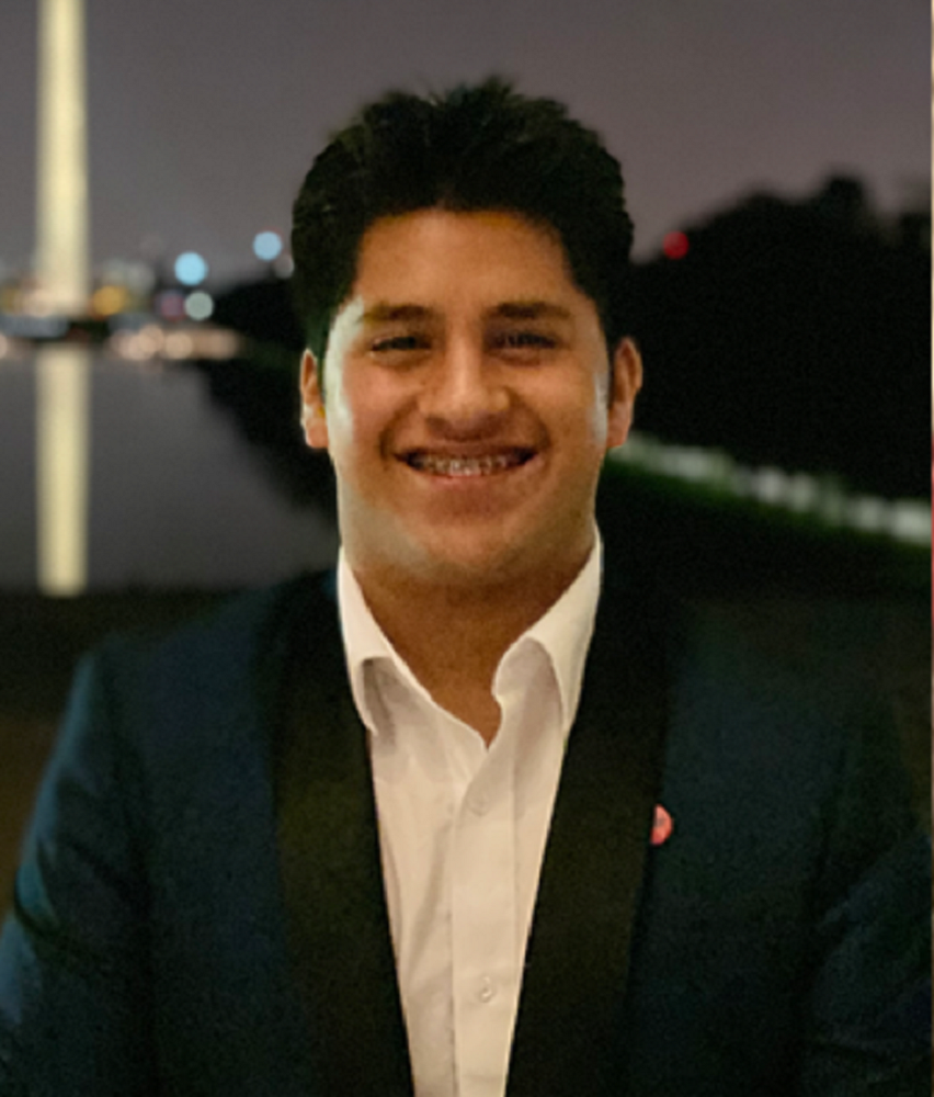 A man dressed in a suit and tie beams at the Washington Monument, with trees and a clear sky in the background.