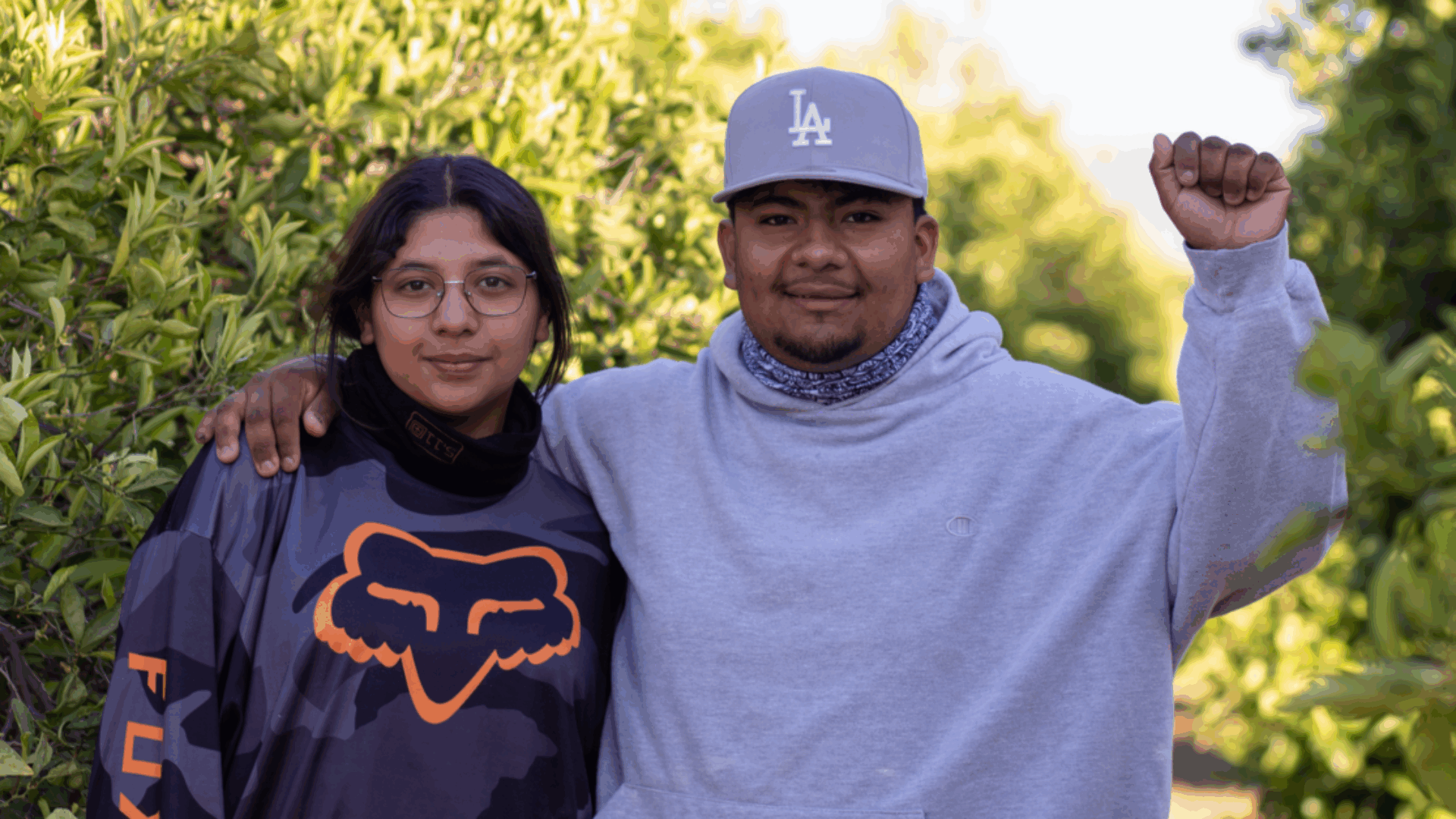 A man and woman stand together in front of lush green trees, smiling at the camera.