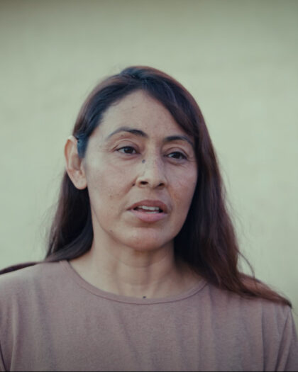 A woman stands outside a house, looking at the camera with a friendly smile, surrounded by a garden.
