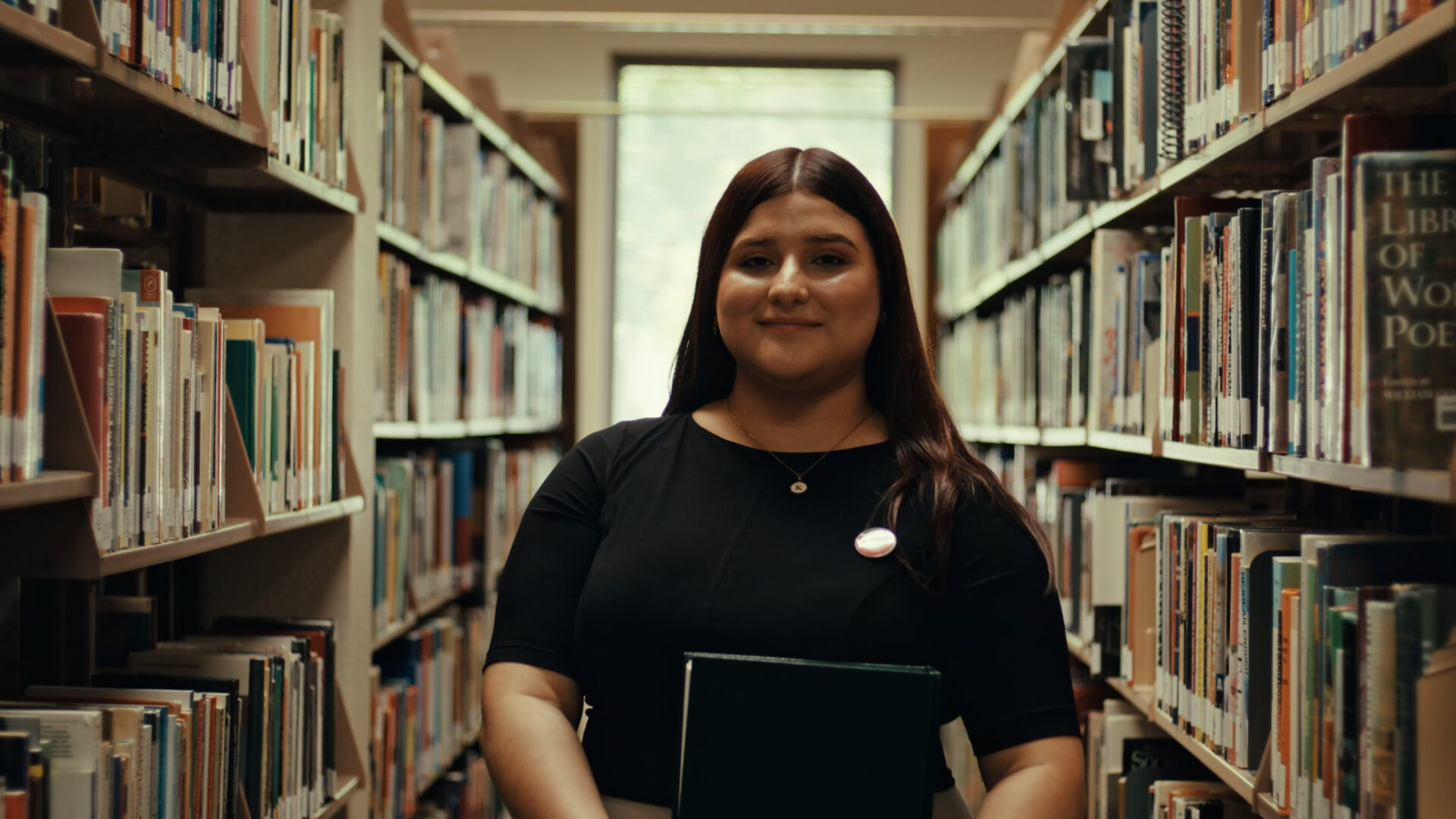 A woman in a library holds a book, surrounded by tall shelves filled with various titles.