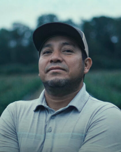 A man standing outdoors in what appears to be a field or garden, with rows of plants visible behind him.