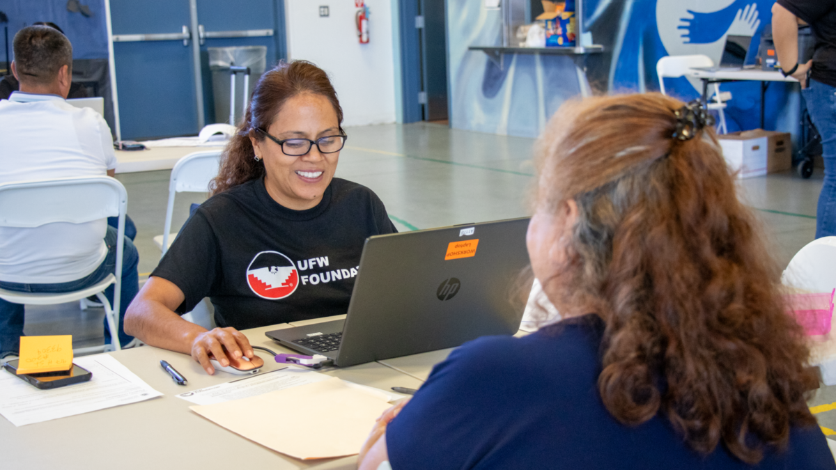 A woman wearing a black UFW Foundation t-shirt smiles while using a laptop and computer mouse at a table with another person.