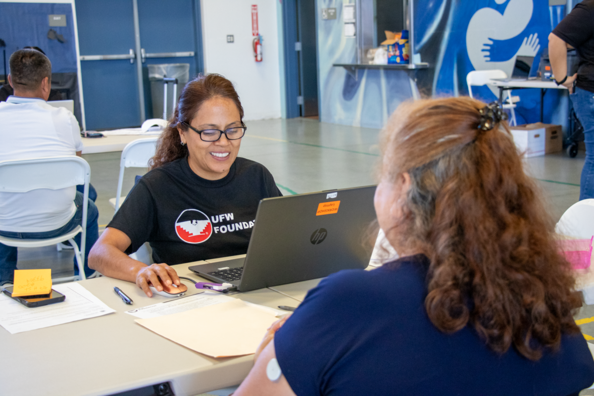 A woman wearing a black UFW Foundation t-shirt smiles while using a laptop and computer mouse at a table with another person.