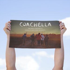 Hands holding a rally towel with farm workers and the text “The real Coachella, home of our farm workers” against a blue sky with clouds.