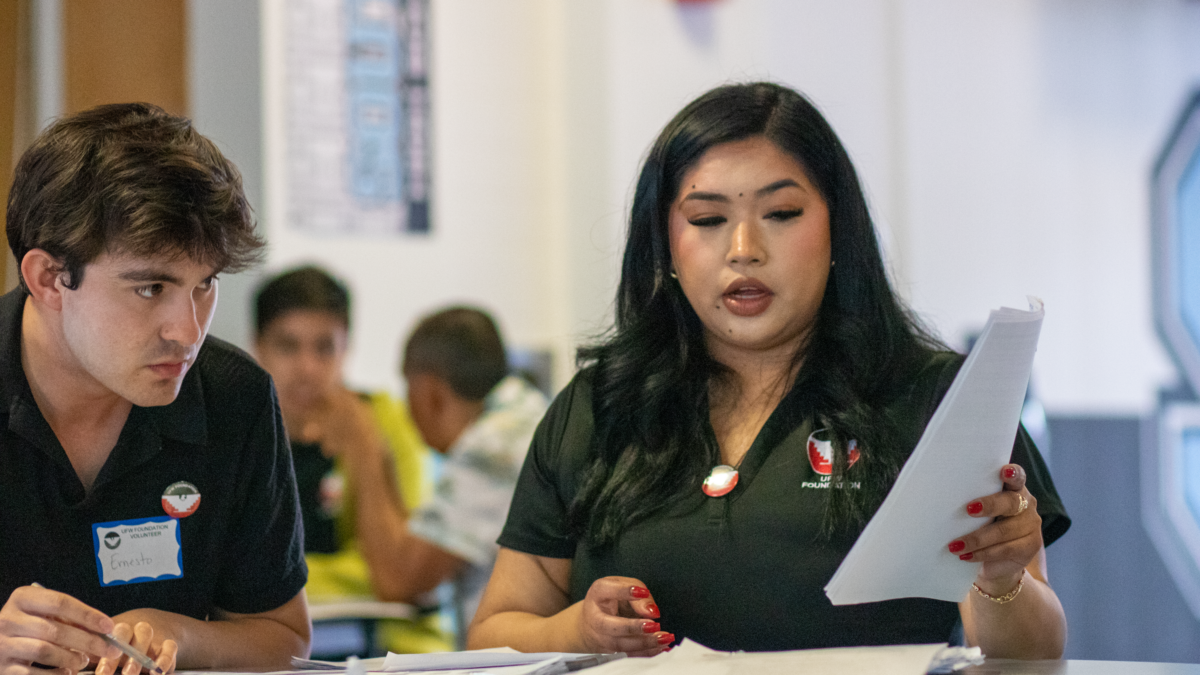 A woman in a black shirt reads documents at a table while a man in a black shirt with a name tag sits to her left.